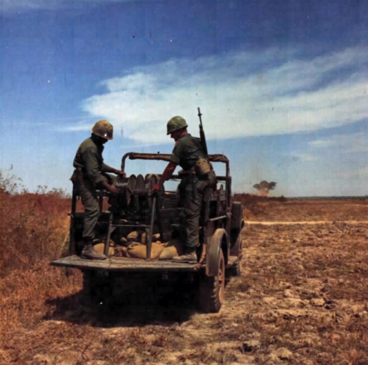 Two soldiers on a moving wire-laying vehicle feeding communication wire out across open ground beside a field.