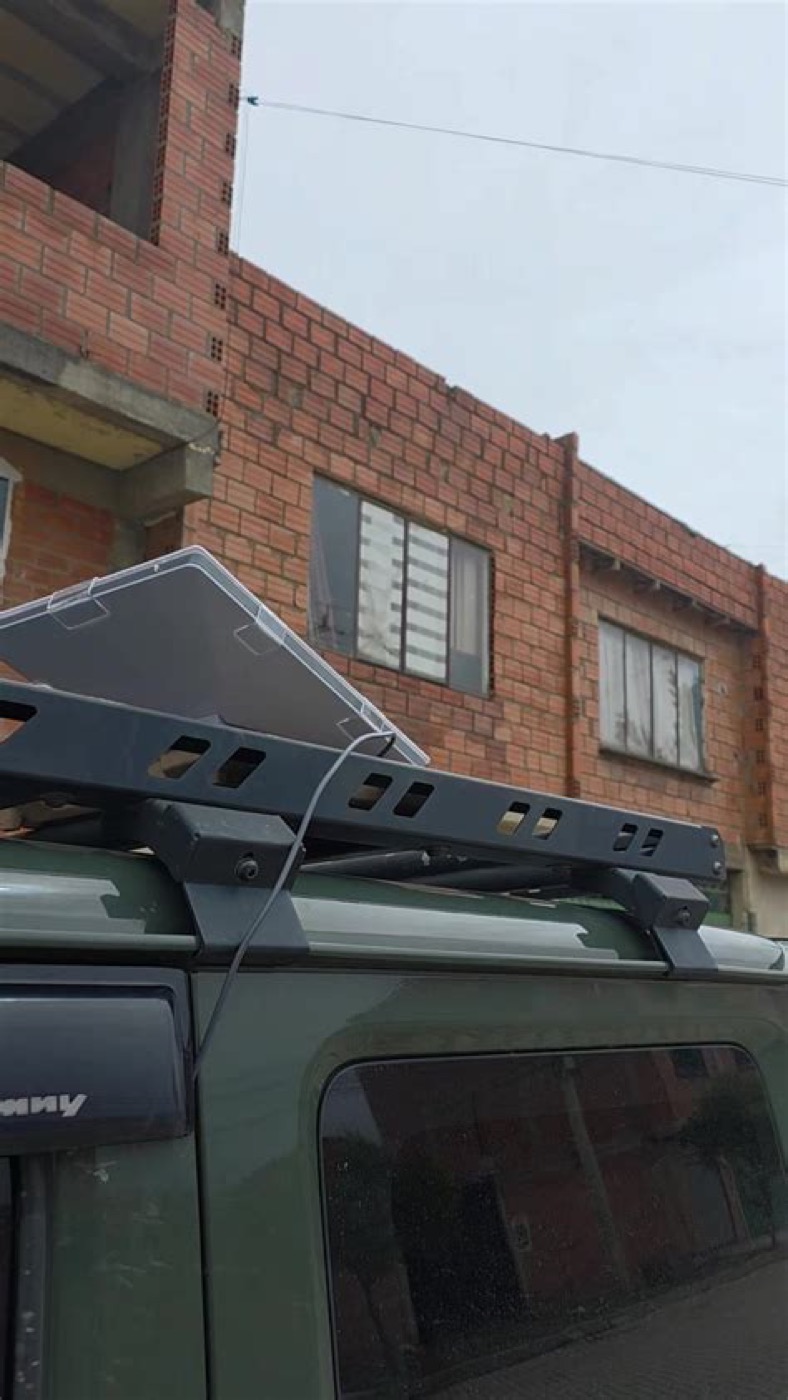 A Starlink flat panel lying on the roof rack of a small four-wheel-drive vehicle parked in a street, its cable running down past the door pillar.