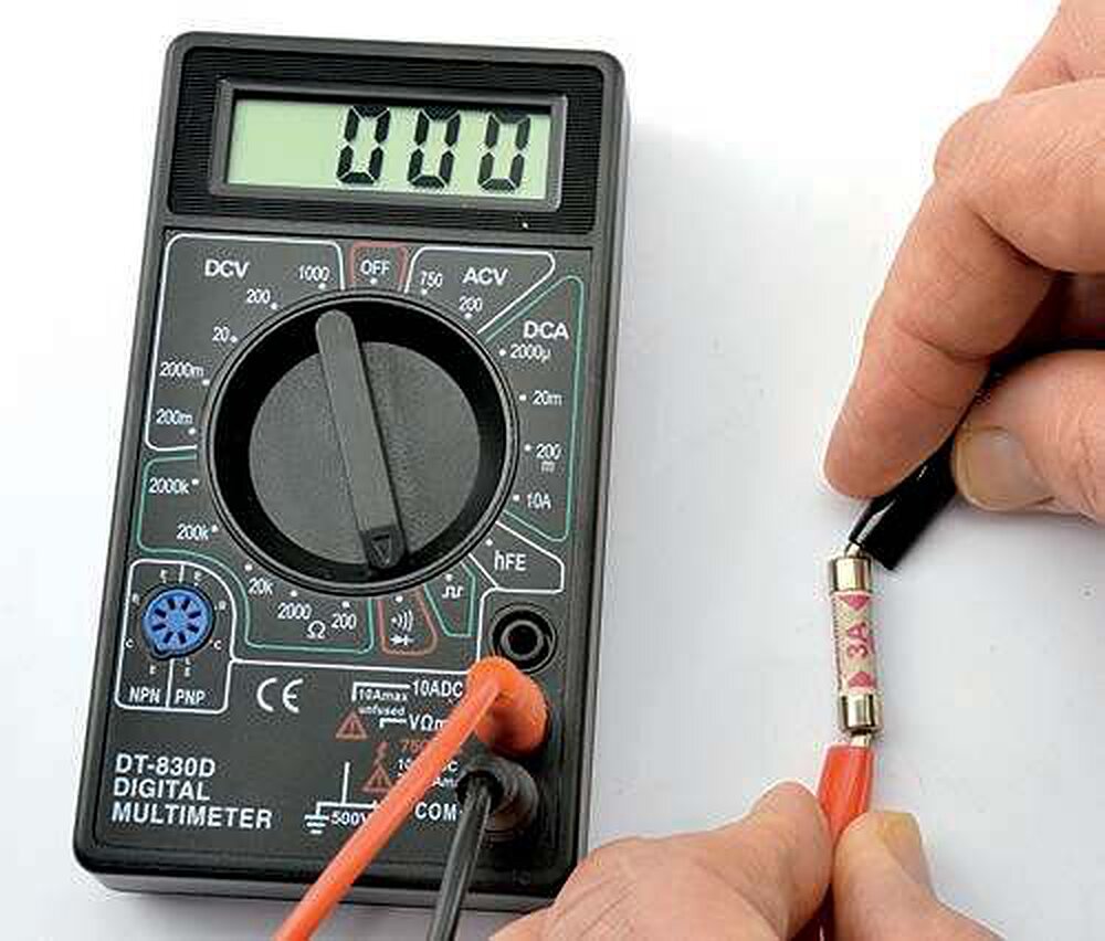 A multimeter set to continuity reading a cartridge fuse held across its probes, display showing zero