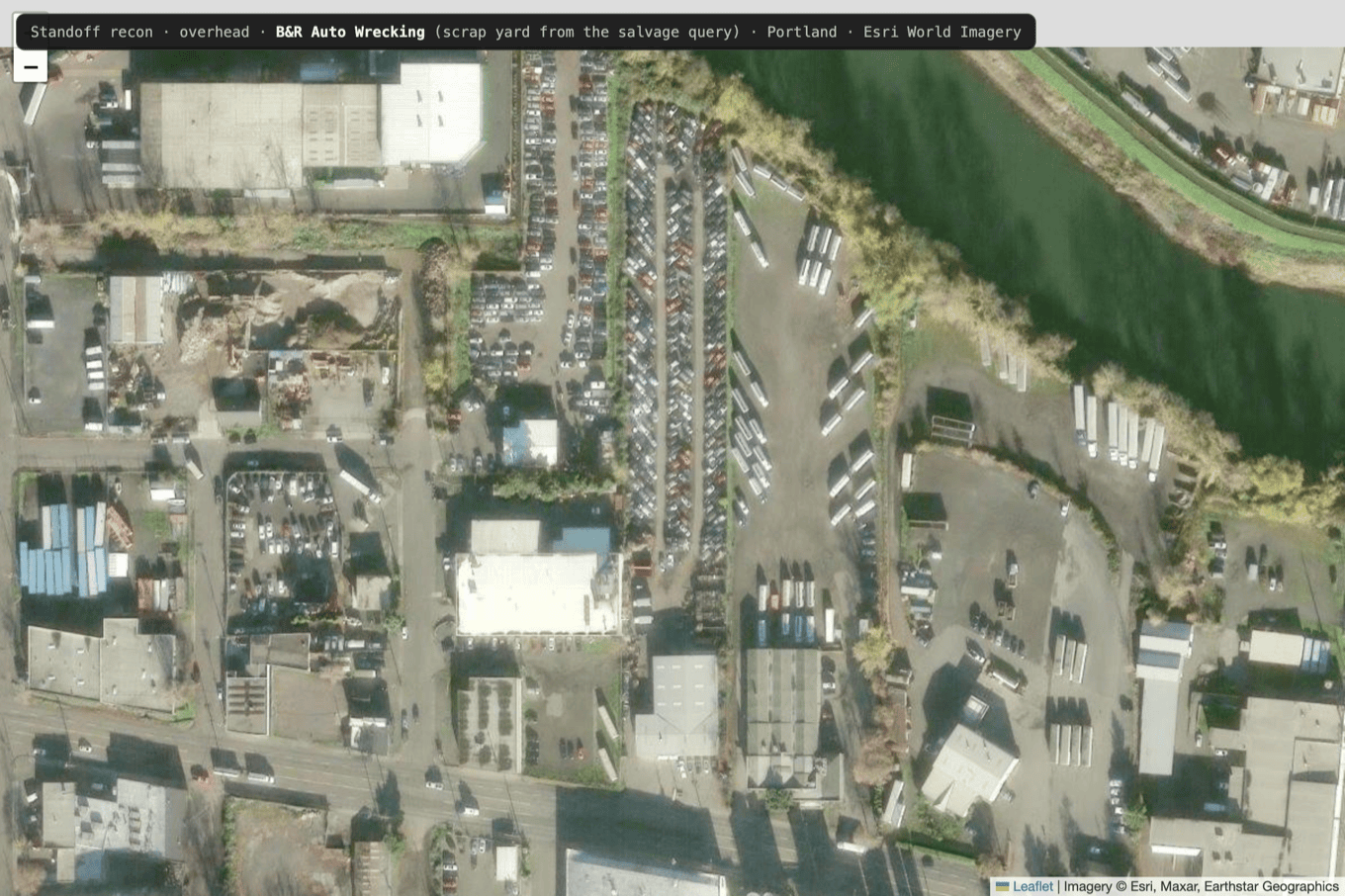 Overhead satellite view of an auto-wrecking scrap yard, with packed rows of wrecked vehicles, an office building, material piles, and the yard perimeter bounded by a river and roads
