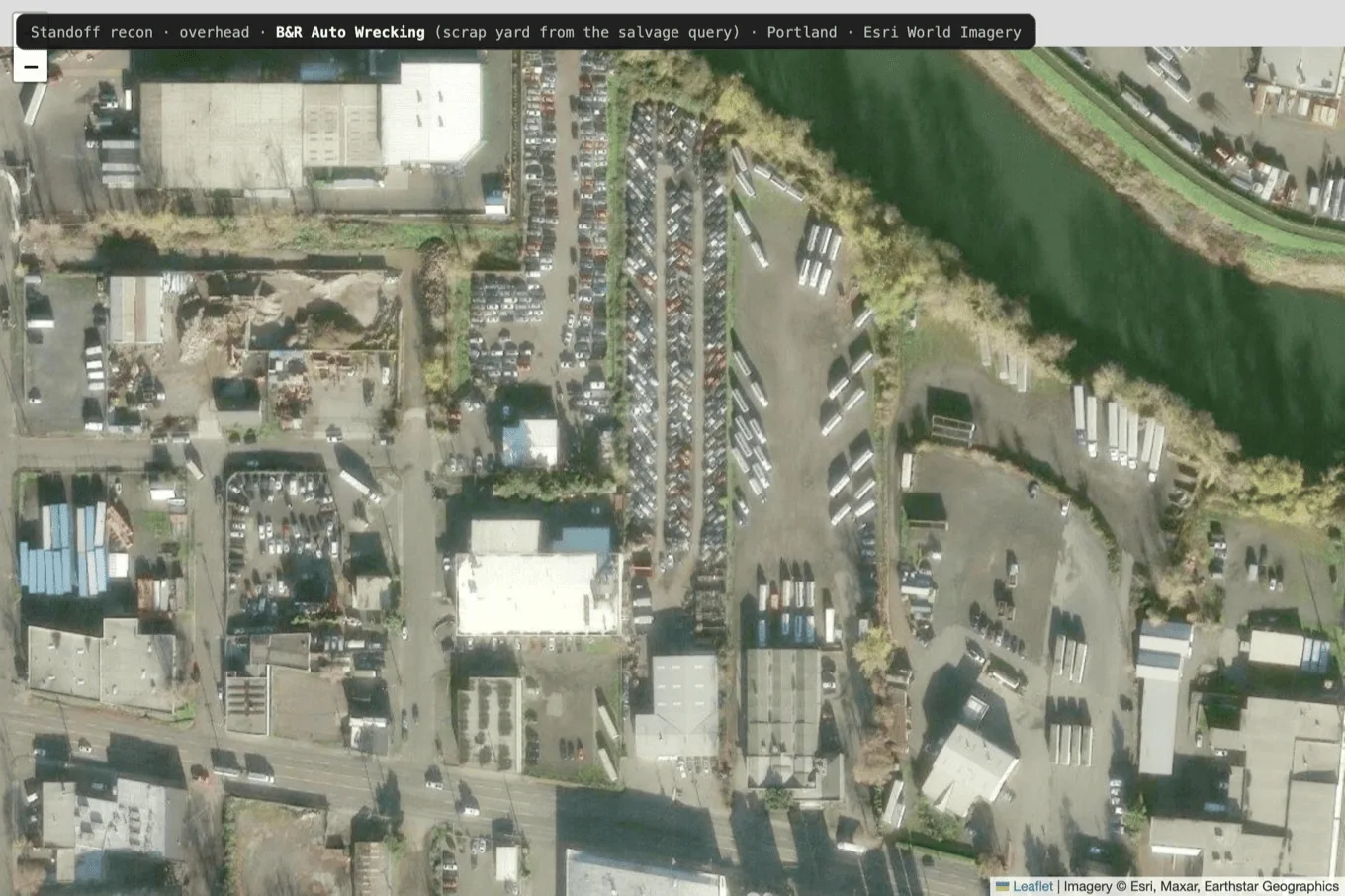 Overhead satellite view of an auto-wrecking scrap yard, with packed rows of wrecked vehicles, an office building, material piles, and the yard perimeter bounded by a river and roads